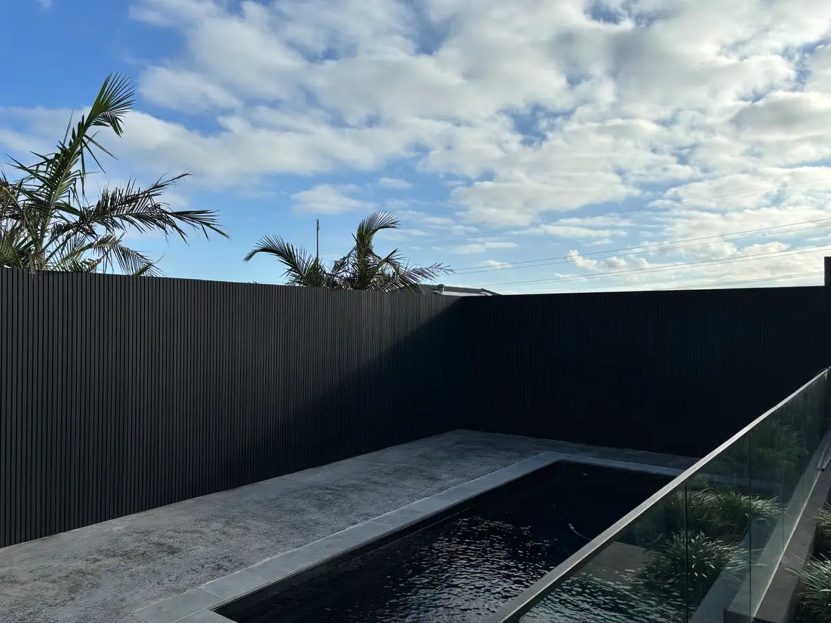 Tall black vertical-slat fence on two sides of a pool with a concrete surround and glass balustrade