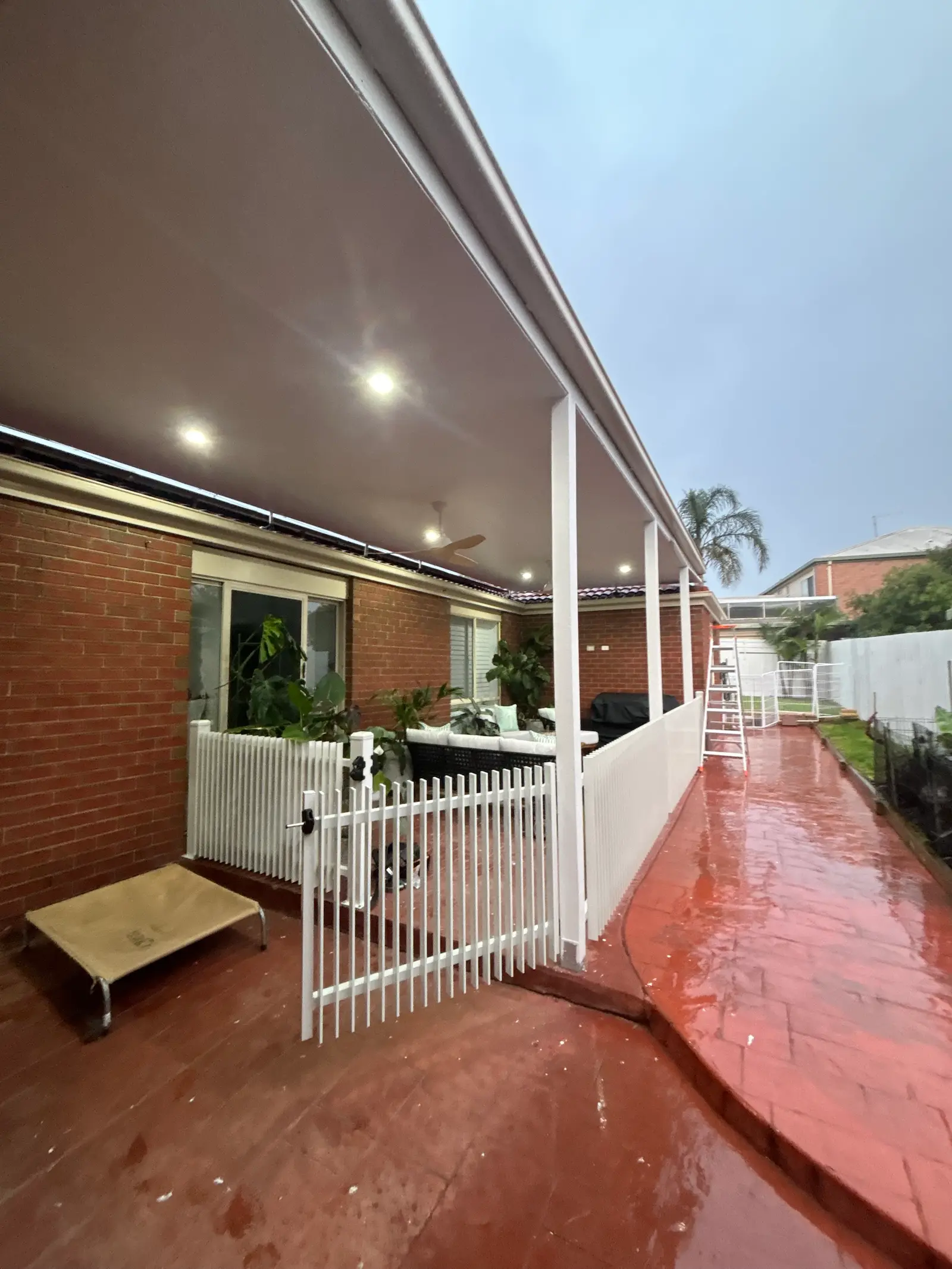 Verandah seen from the side path on a wet day, with the white picket gate, downlights and ceiling fans
