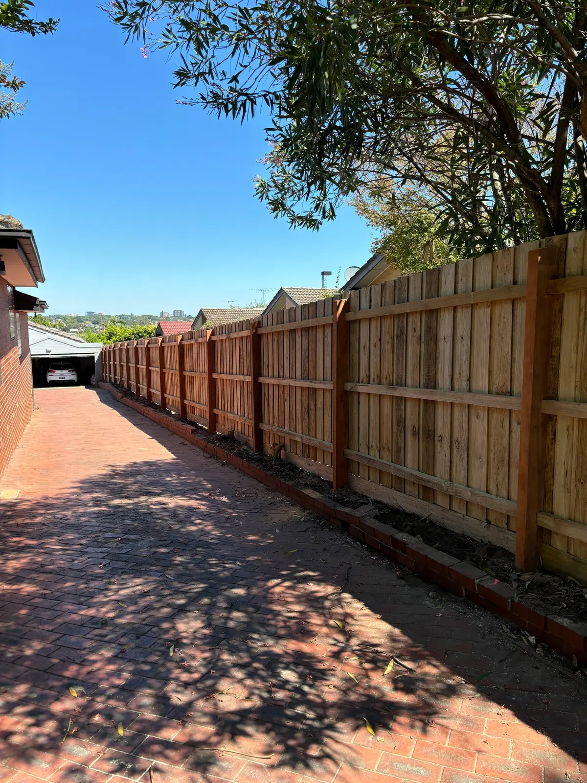 Treated pine paling fence with exposed posts and rails running alongside a brick-paved driveway towards a garage