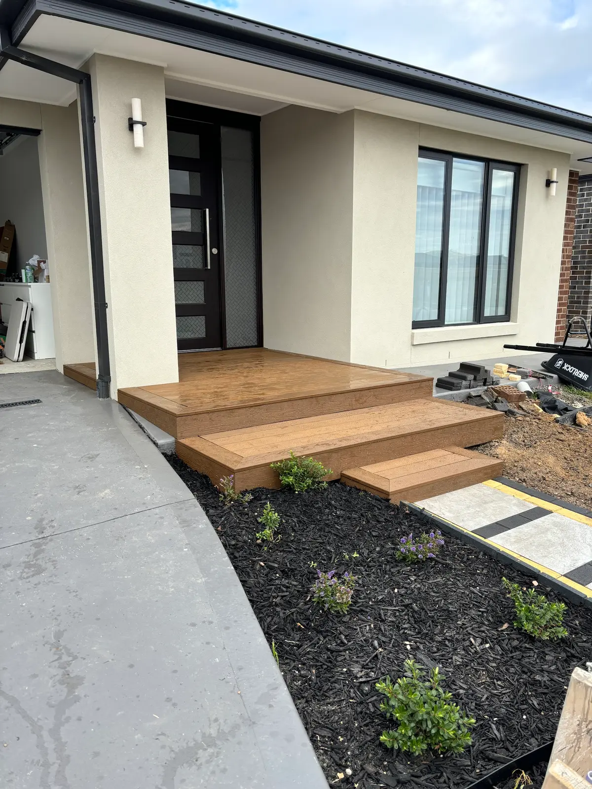 Low timber-look entry deck with picture-framed edges and two wide steps at the front door of a rendered home, beside a mulched garden bed