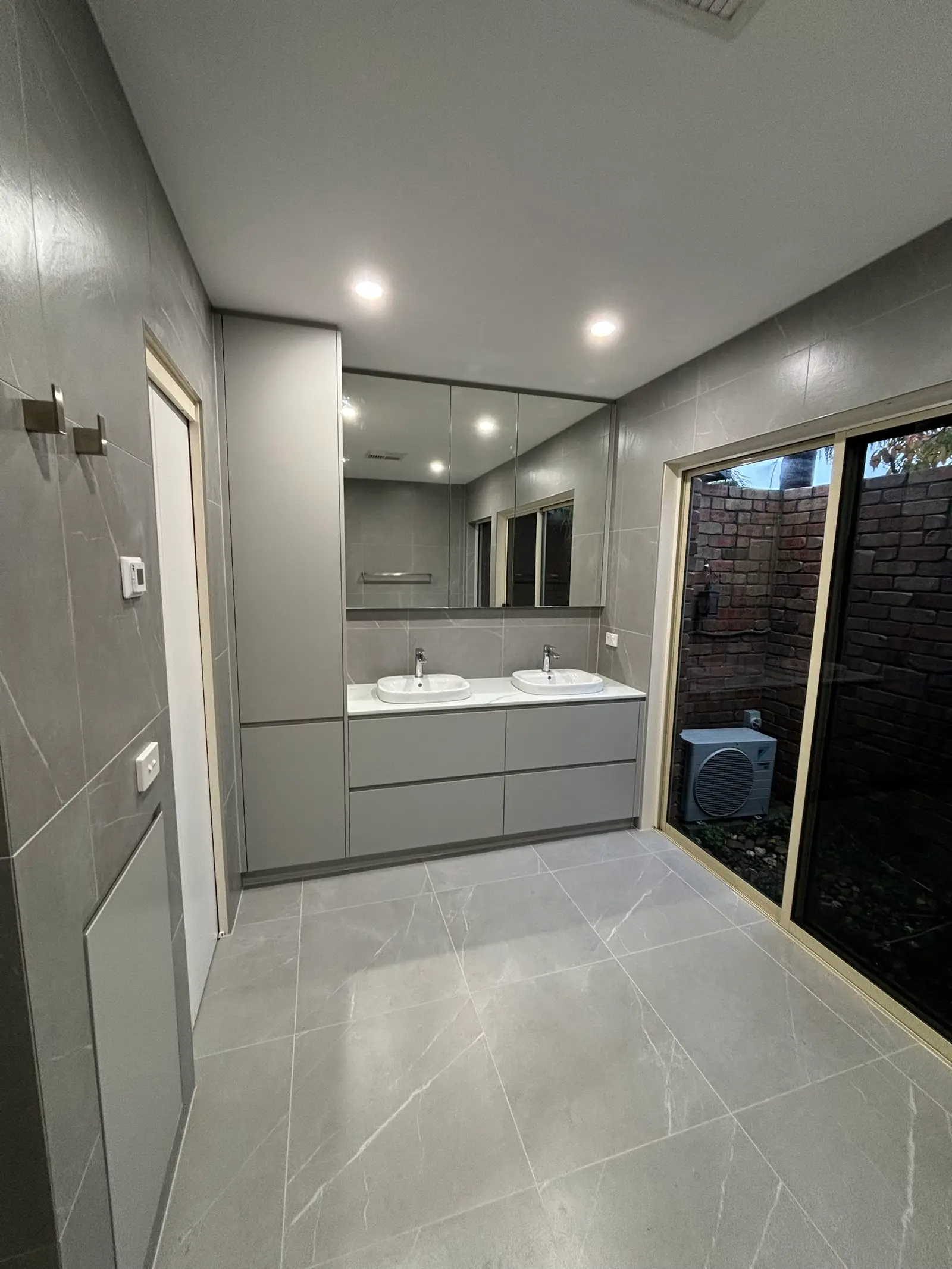 Renovated grey bathroom with a double vanity, floor-to-ceiling cabinet, mirrored cabinet and large-format tiles, beside a sliding door to a courtyard