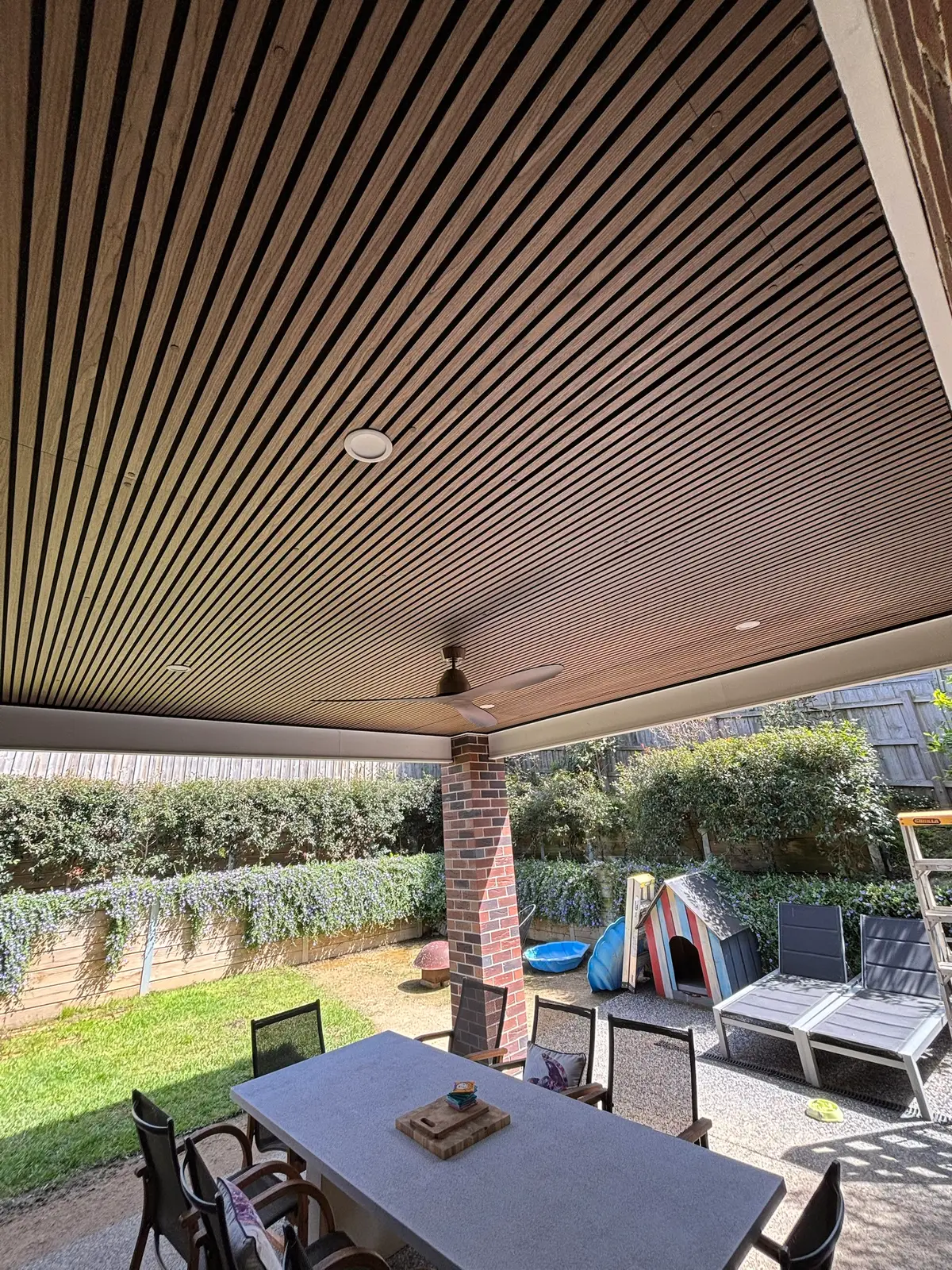 Timber-look slat ceiling over a covered alfresco with a ceiling fan, downlights, brick pier and outdoor dining table