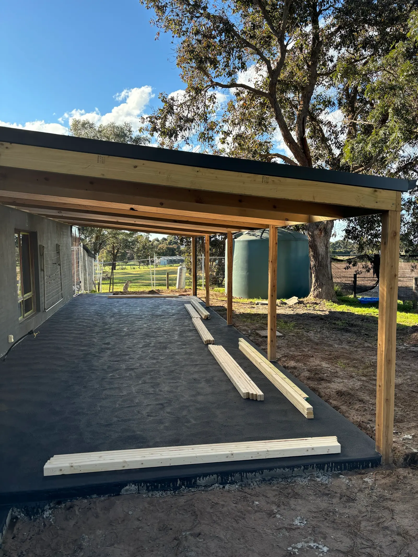 The same structure during construction: timber beams and rafters under steel roofing on posts over the new slab, before the walls were clad