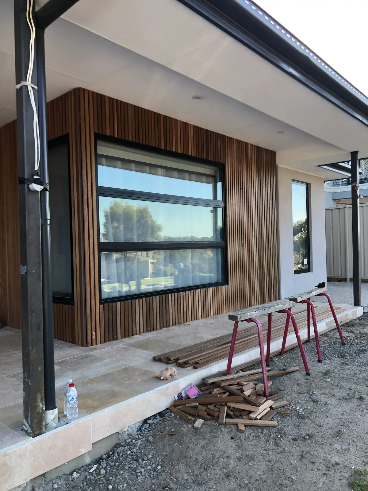 Vertical hardwood battens being installed around a bay window with black frames under a wide eave, with cut battens on sawhorses